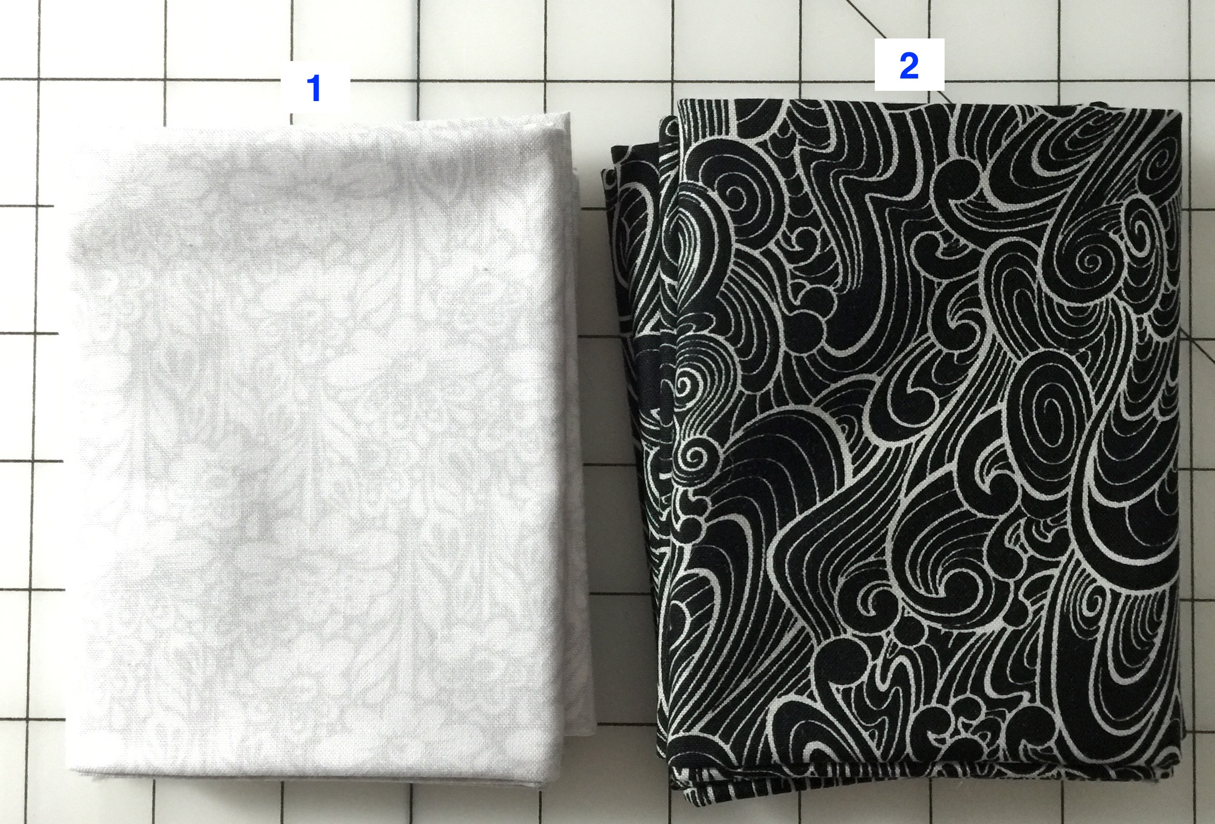 fabrics1-2 Two folded pieces of fabric, one is white with light grey florals printed and one is black with silver swirly designs on it.